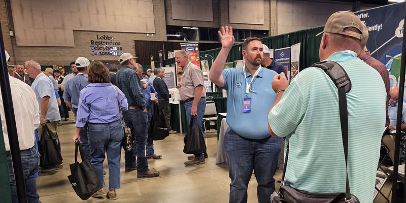 A crowded trade show floor, one attendee waving at the camera