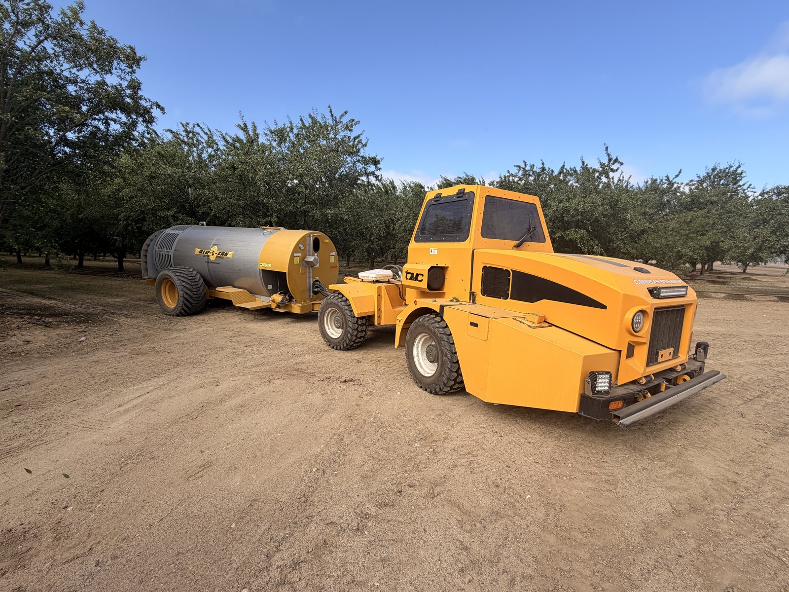 A modern yellow orchard tractor with a spray tank parked in a California almond orchard