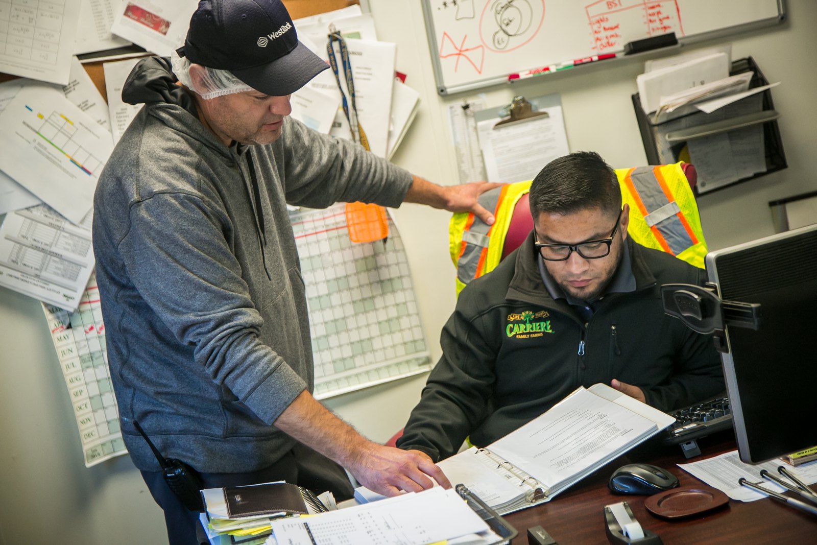 A farm supervisor and a worker in a high-visibility vest review a binder of safety documentation together at an office desk
